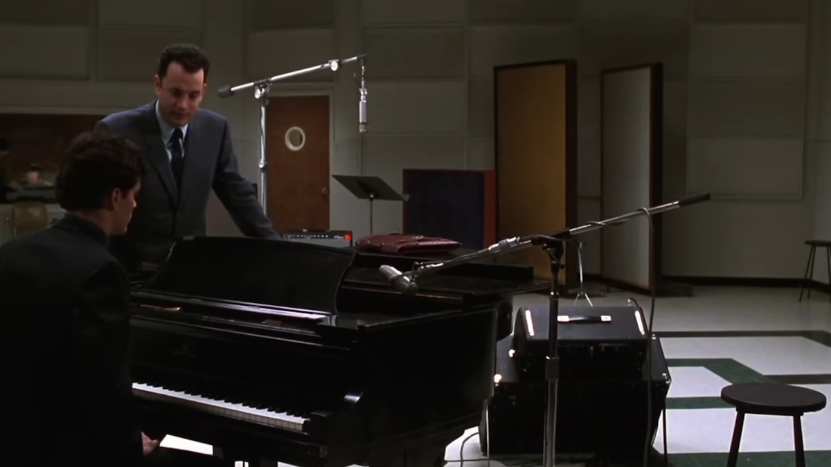 Tom Hanks stojí v hudebním nahrávacím studiu a opírá se o černý klavír ve filmu That Thing You Do!