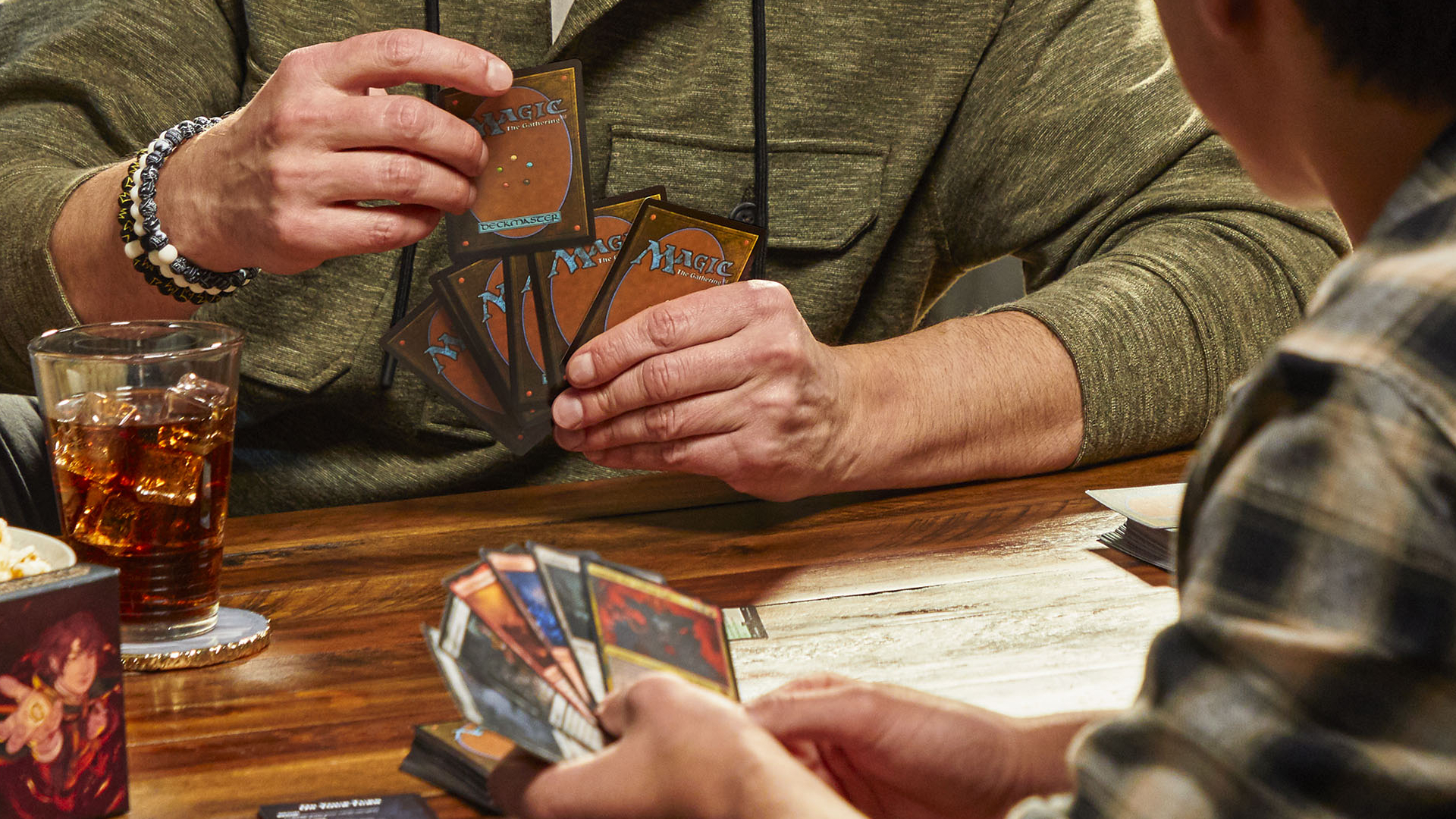 Duas pessoas seguram cartas de Magic: The Gathering, sentadas em frente uma da outra numa mesa