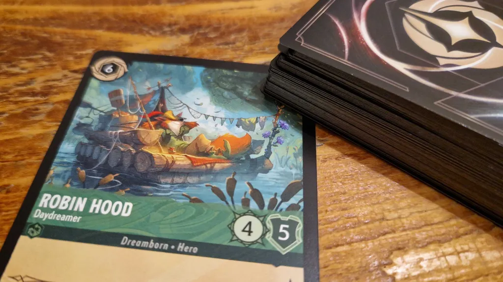 Robin Hood odpočívá na voru na obrázku z karet Disney Lorcana Into the Inklands, položeném na dřevěném stole vedle balíčku karet.